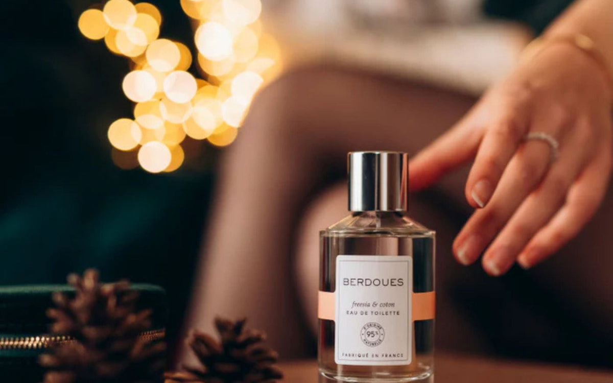 Flacon d’eau de toilette Berdoues posé sur une table avec une main féminine s’en approchant, ambiance douce et lumineuse