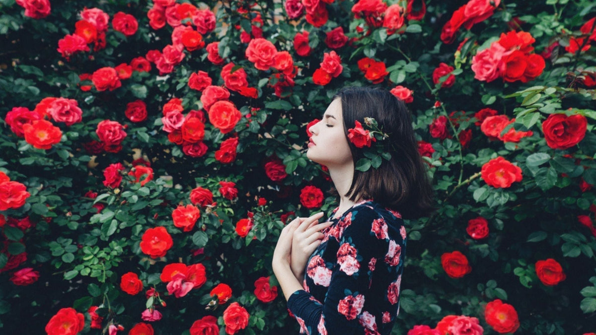 Femme respirant le parfum des roses dans un jardin luxuriant, illustrant les ingrédients rares et précieux en parfumerie.