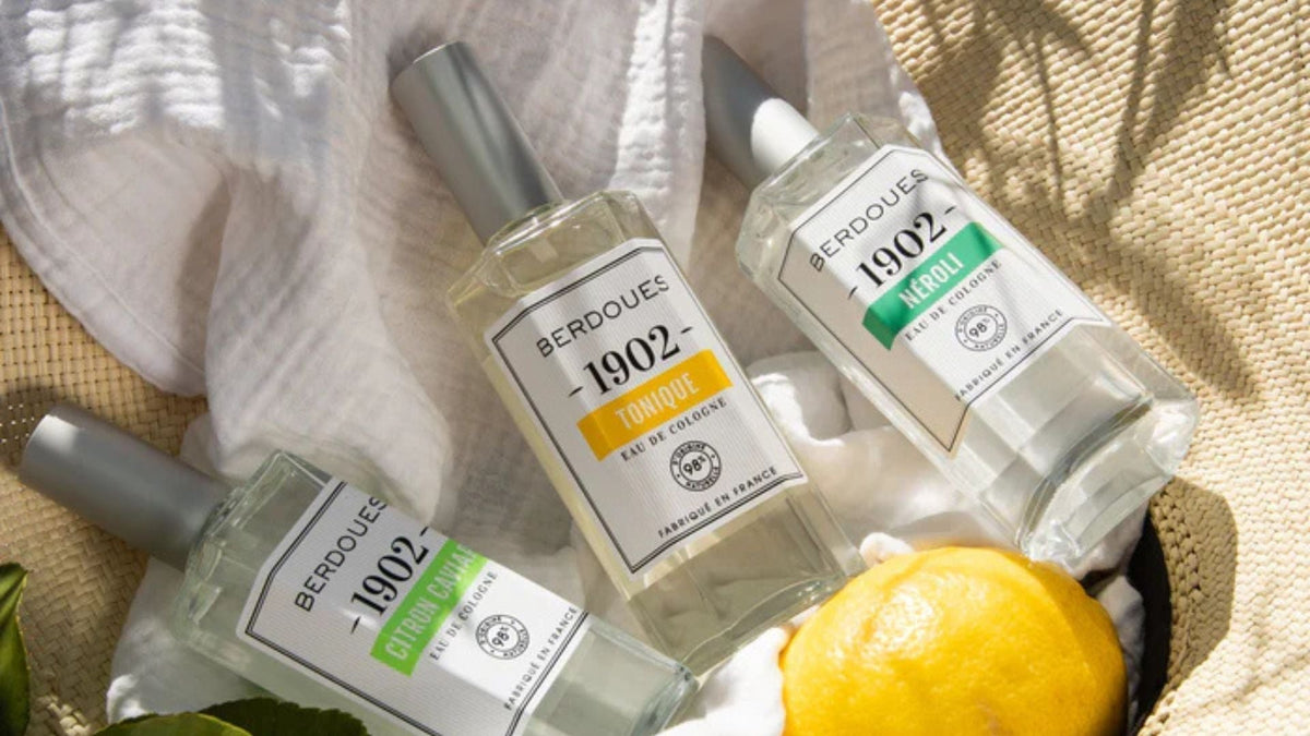 Flacons d’eau de Cologne Berdoues 1902 posés sur un tissu blanc avec un citron, illustrant le renouveau des parfums frais et authentiques chez les jeunes générations.